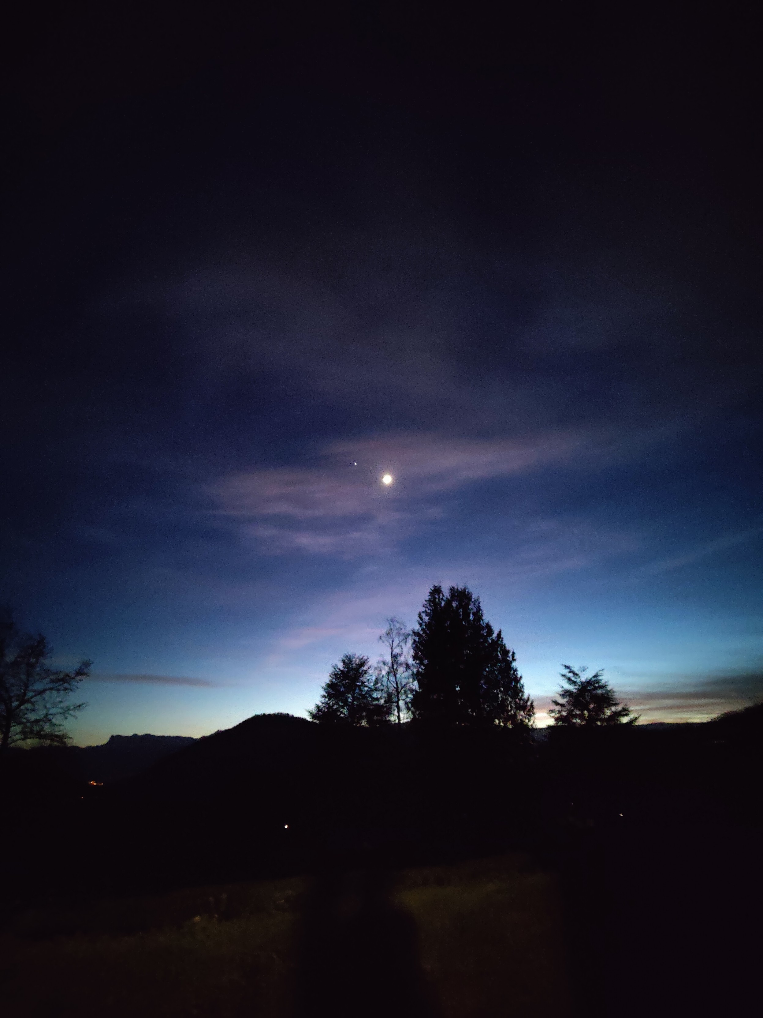 Les dernières lumières du soleil sont en train de disparaître à l'horizon derrière les montagnes. L'ombre des arbres se détache devant un dégradé de bleu clair à bleu nuit. Dans le ciel, un croissant de lune et un point lumineux (étoile, planète?) brillent côte à côte derrière un petit voile nuageux.