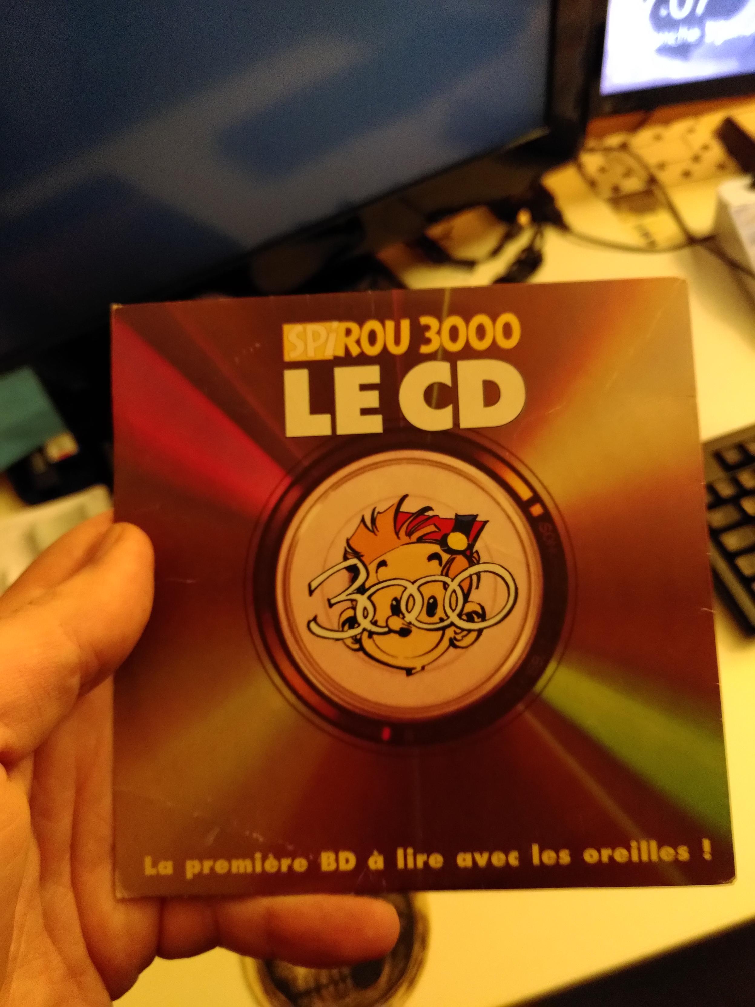 Photo de ma main tenant le CD de "Spirou 3000". On y voit une tête de Spirou avec des lunettes qui forment le chiffre 3000 au centre. En bas, un slogan : La première BD à lire avec les oreilles !